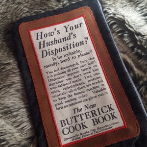 May include: A brown leather patch with a white background and red text. The text reads "How's Your Husband's Disposition? Is he irritable, moody, hard to please?" and continues with a paragraph about the importance of a well-balanced diet for a husband's disposition. The patch also includes the text "The New Butterick Cook Book" and "Sponsored by Martha Van Rensselaer, Home Making Editor of The Delineator".