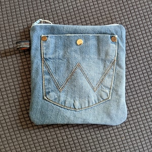 Puede incluir: Una bolsa de cremallera de mezclilla azul con un bolsillo grande en la parte delantera. El bolsillo tiene un detalle de costura en forma de W cl&aacute;sico y dos remaches de color dorado.