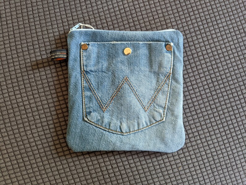 Puede incluir: Una bolsa de cremallera de mezclilla azul con un bolsillo grande en la parte delantera. El bolsillo tiene un detalle de costura en forma de W cl&aacute;sico y dos remaches de color dorado.