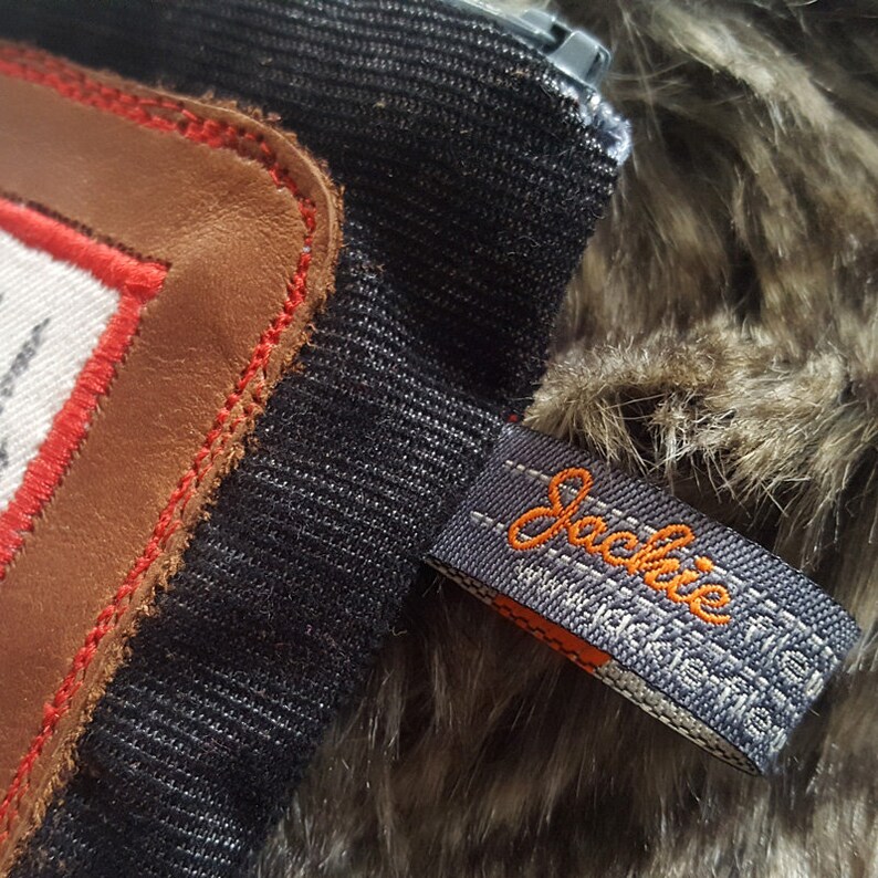 May include: A black zippered pouch with a brown leather patch and red stitching. The pouch has a gray fabric tag with the word "Jackie" in orange thread.
