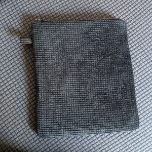 May include: A gray zippered pouch with a textured fabric. The pouch is lying on a gray and white patterned surface.