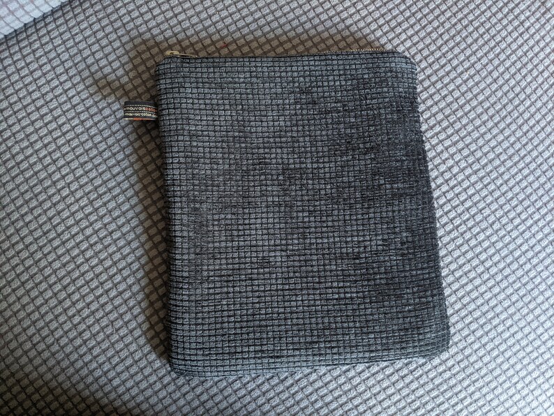 May include: A gray zippered pouch with a textured fabric. The pouch is lying on a gray and white patterned surface.