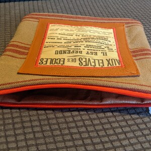 May include: A brown and tan striped fabric zippered pouch with a brown leather flap. The flap has a vintage newspaper clipping sewn onto it that reads "Aux Eleves des Ecoles Il Est Defendue". The pouch has a red zipper and a price tag that reads "2.25e".