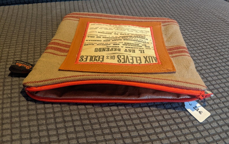 May include: A brown and tan striped fabric zippered pouch with a brown leather flap. The flap has a vintage newspaper clipping sewn onto it that reads "Aux Eleves des Ecoles Il Est Defendue". The pouch has a red zipper and a price tag that reads "2.25e".
