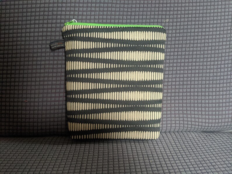 May include: A black and beige zippered pouch with a green zipper. The pouch has a repeating pattern of beige lines on a black background.