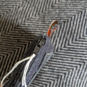 May include: A black zippered pouch with a white string attached to the zipper pull. The pouch is made of a textured fabric and has a black zipper.