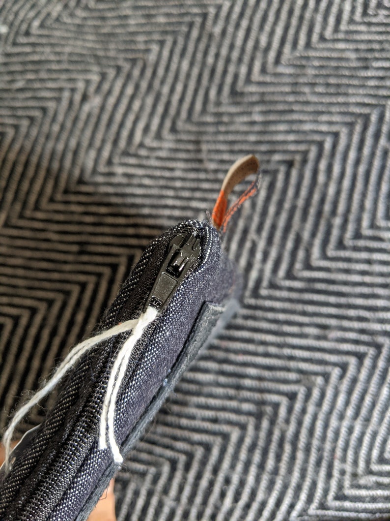 May include: A black zippered pouch with a white string attached to the zipper pull. The pouch is made of a textured fabric and has a black zipper.