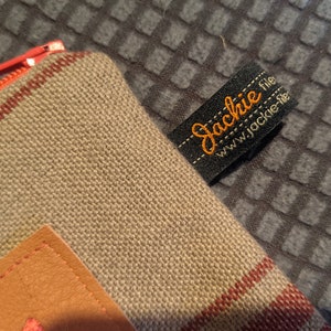 May include: A beige canvas zippered pouch with a brown leather patch and red stripes. The pouch has a black label with the text "Jackie file www.jackie-file" in orange.