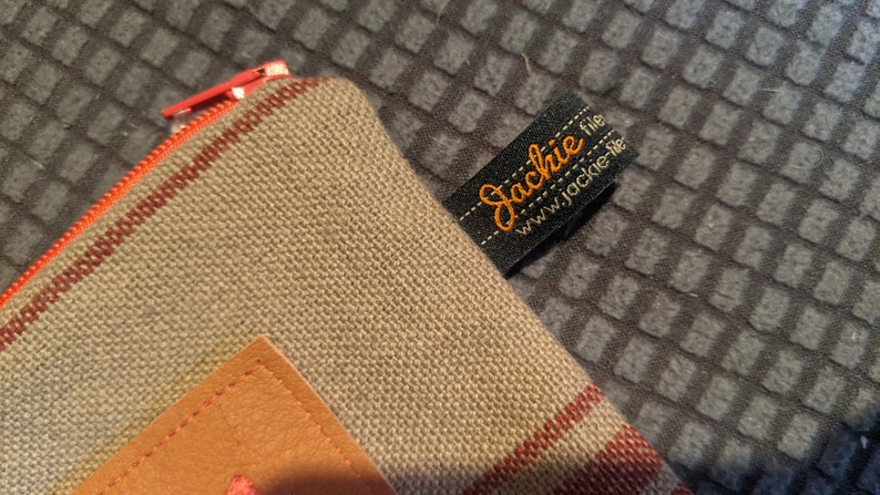 May include: A beige canvas zippered pouch with a brown leather patch and red stripes. The pouch has a black label with the text "Jackie file www.jackie-file" in orange.