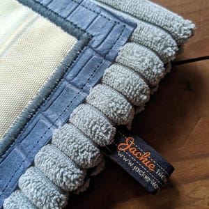 K&ouml;nnte beinhalten: Ein hellblauer und wei&szlig; gestreifter Stoff mit einem blauen geh&auml;kelten Besatz und einem blauen lederartigen Rand. Der Stoff hat ein Etikett mit der Aufschrift "Jackie Fisher" und "www.jackie-fisher.com".