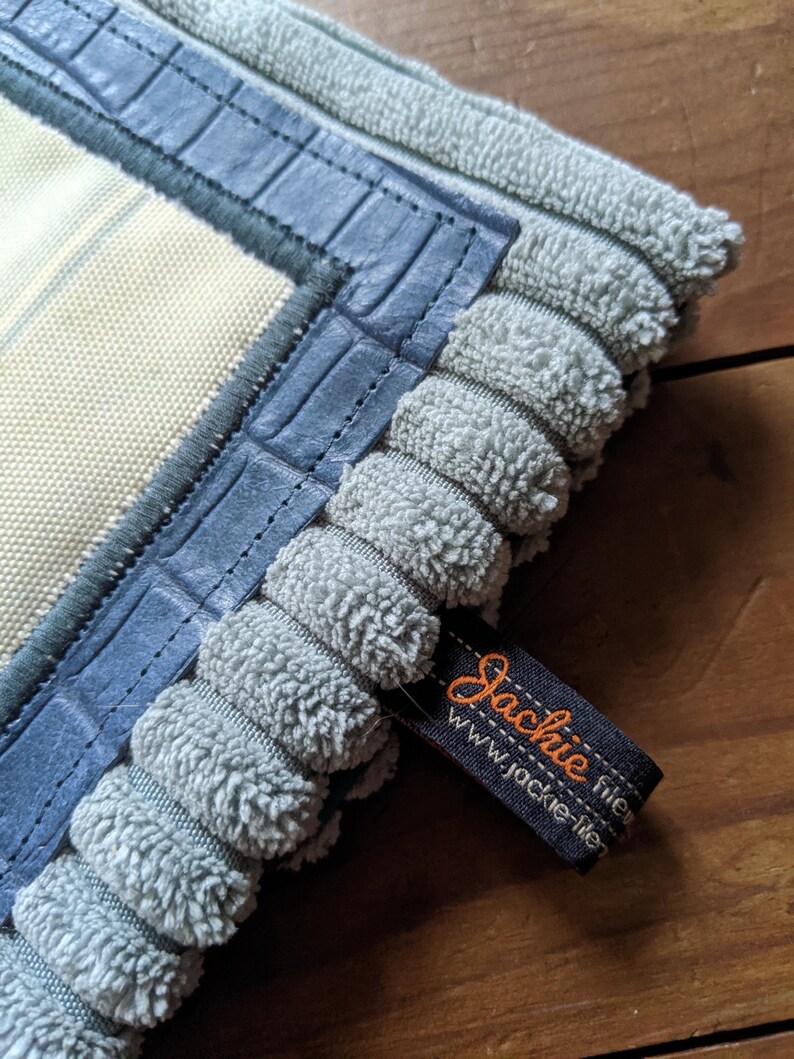 K&ouml;nnte beinhalten: Ein hellblauer und wei&szlig; gestreifter Stoff mit einem blauen geh&auml;kelten Besatz und einem blauen lederartigen Rand. Der Stoff hat ein Etikett mit der Aufschrift "Jackie Fisher" und "www.jackie-fisher.com".