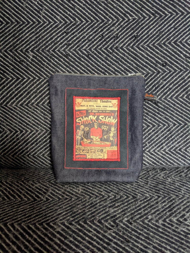 Puede incluir: Una bolsa de cremallera de mezclilla azul con un p&oacute;ster vintage rojo y blanco con el texto "Dr. Brundell's Shock Show".