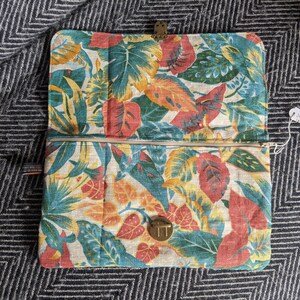 Pu&ograve; includere: Una pochette con zip con stampa floreale che presenta foglie rosse, gialle e blu su uno sfondo bianco. La pochette ha una zip dorata e due chiusure dorate.