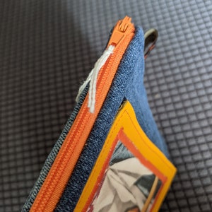 May include: A blue denim zippered pouch with an orange zipper and a yellow border around a colorful image inside.