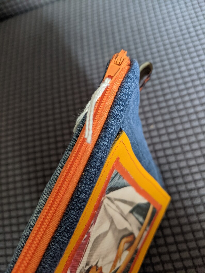 May include: A blue denim zippered pouch with an orange zipper and a yellow border around a colorful image inside.