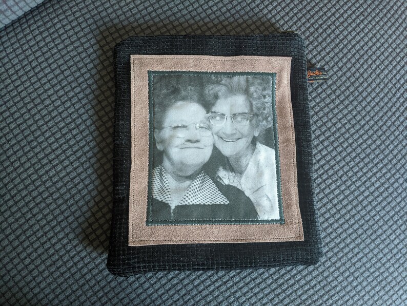 May include: A black fabric pouch with a brown suede frame and a black and white photo of two women smiling. The photo is framed with a green border.