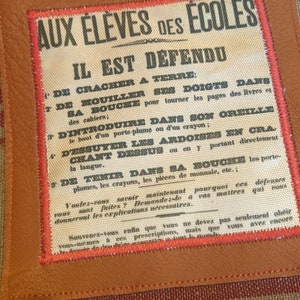 May include: A vintage-style sign with red and black text on a white background. The sign reads "Aux Eleves des Ecoles Il Est Defendu" and lists a series of rules for students. The sign is framed in brown leather with red stitching.