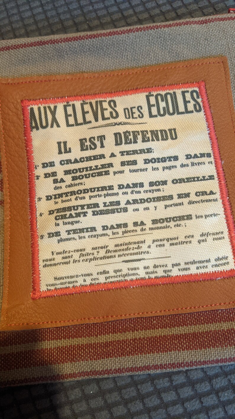 May include: A vintage-style sign with red and black text on a white background. The sign reads "Aux Eleves des Ecoles Il Est Defendu" and lists a series of rules for students. The sign is framed in brown leather with red stitching.