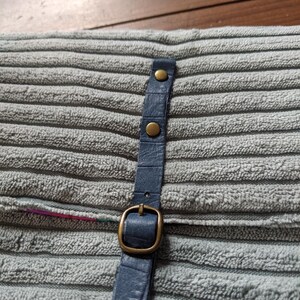 K&ouml;nnte beinhalten: Ein blaues Lederband mit einer Messing-Schnalle und Nieten. Das Band ist an einer grauen Stoffoberfl&auml;che befestigt.