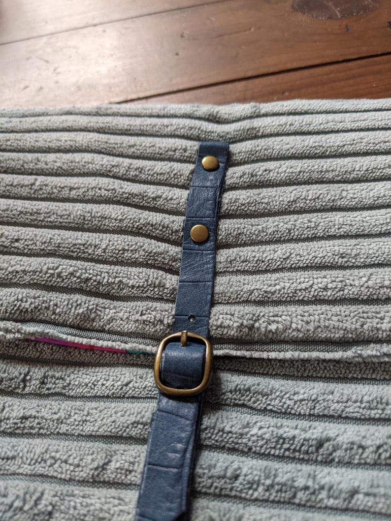 K&ouml;nnte beinhalten: Ein blaues Lederband mit einer Messing-Schnalle und Nieten. Das Band ist an einer grauen Stoffoberfl&auml;che befestigt.