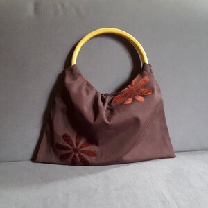 以下が含まれることがあります： 木製のハンドルと刺繍された茶色の花が付いた茶色の布製のトートバッグ。