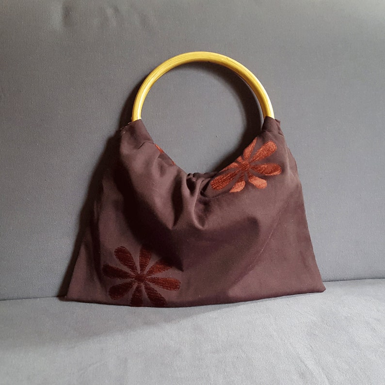 以下が含まれることがあります： 木製のハンドルと刺繍された茶色の花が付いた茶色の布製のトートバッグ。