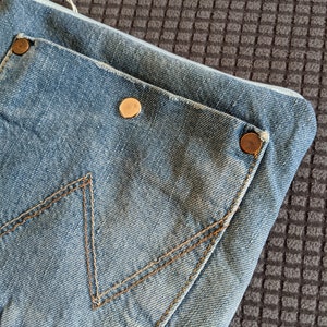 Puede incluir: Un clutch de mezclilla azul con cierre de cremallera y un bolsillo grande en la parte delantera. El bolsillo tiene un detalle de costura marr&oacute;n y dos botones met&aacute;licos dorados.