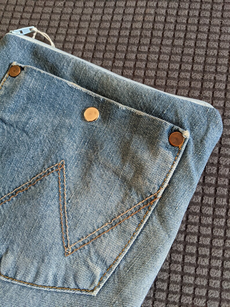 Puede incluir: Un clutch de mezclilla azul con cierre de cremallera y un bolsillo grande en la parte delantera. El bolsillo tiene un detalle de costura marr&oacute;n y dos botones met&aacute;licos dorados.