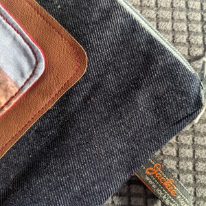 K&ouml;nnte beinhalten: Eine dunkelblaue Jeans-Rei&szlig;verschlusstasche mit einem braunen Leder-Patch und roten N&auml;hten. Die Tasche hat ein Etikett mit der Aufschrift "Sockie" und einer Website-Adresse.