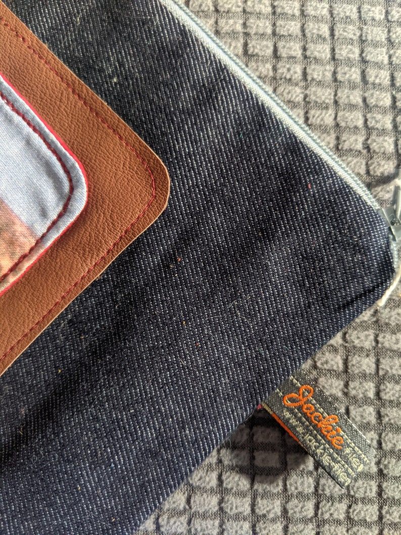 K&ouml;nnte beinhalten: Eine dunkelblaue Jeans-Rei&szlig;verschlusstasche mit einem braunen Leder-Patch und roten N&auml;hten. Die Tasche hat ein Etikett mit der Aufschrift "Sockie" und einer Website-Adresse.