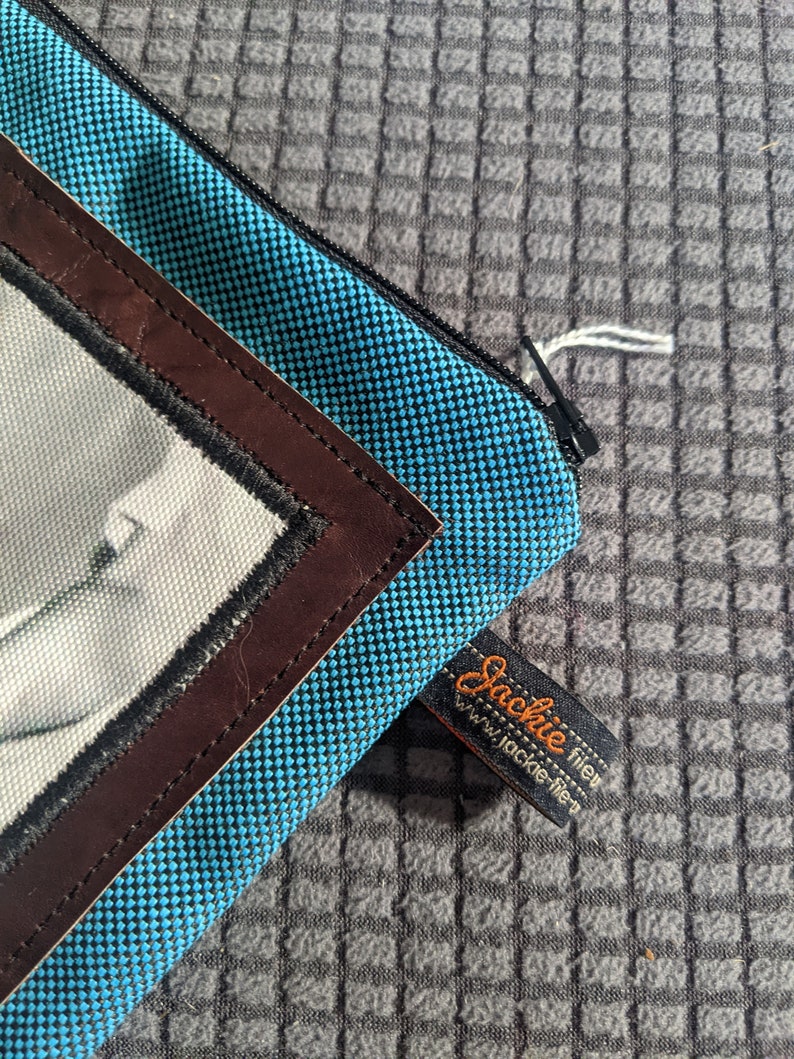 May include: A blue zippered pouch with a brown leather trim and a white and black photo insert. The pouch has a black tag that reads "Jackie tien" and "www.jackie-tien.com".