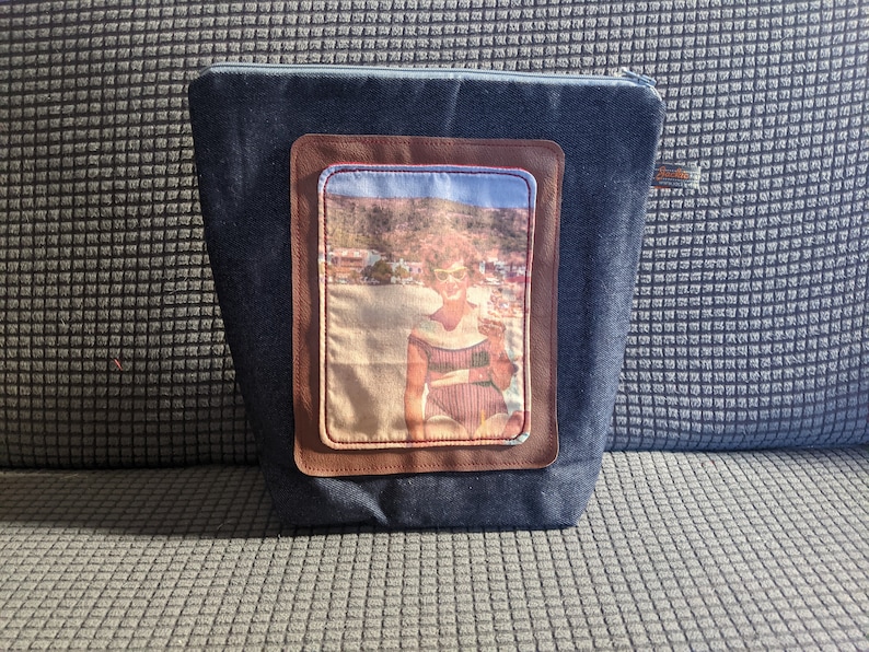 K&ouml;nnte beinhalten: Eine Rei&szlig;verschlusstasche aus Denim mit einem braunen Lederpatch, der ein Vintage-Foto einer Frau im Badeanzug am Strand zeigt.