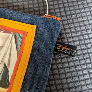 May include: A blue denim zippered pouch with a yellow and orange trim. The pouch has a vintage image of a woman in a white dress. The pouch has a black label with the text "Jackie fileu www.jackie fileu.com".