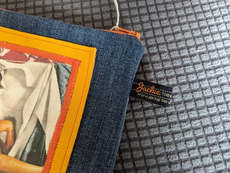 May include: A blue denim zippered pouch with a yellow and orange trim. The pouch has a vintage image of a woman in a white dress. The pouch has a black label with the text "Jackie fileu www.jackie fileu.com".