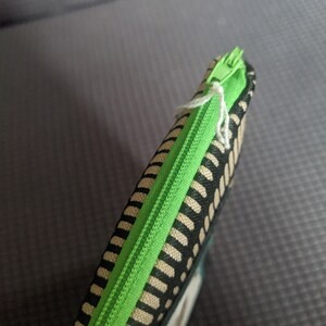 May include: A green zipper runs along the edge of a black and beige striped fabric pouch.