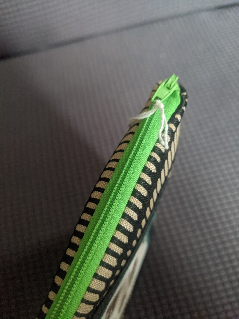 May include: A green zipper runs along the edge of a black and beige striped fabric pouch.