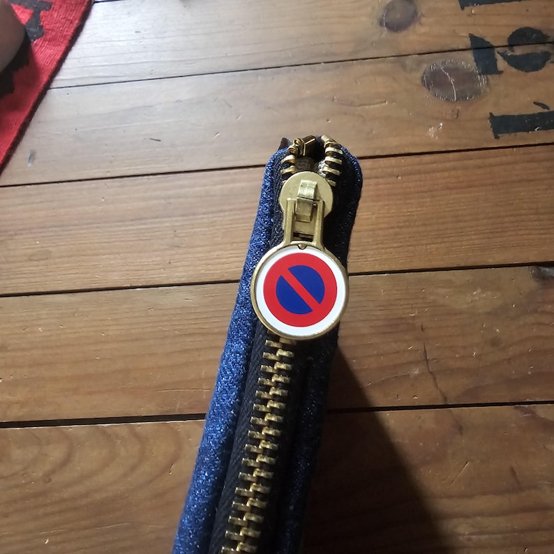 May include: A blue denim wallet with a gold zipper and a round, red, white, and blue no parking sign charm. The wallet is partially open, revealing the black interior. The zipper is a prominent feature.