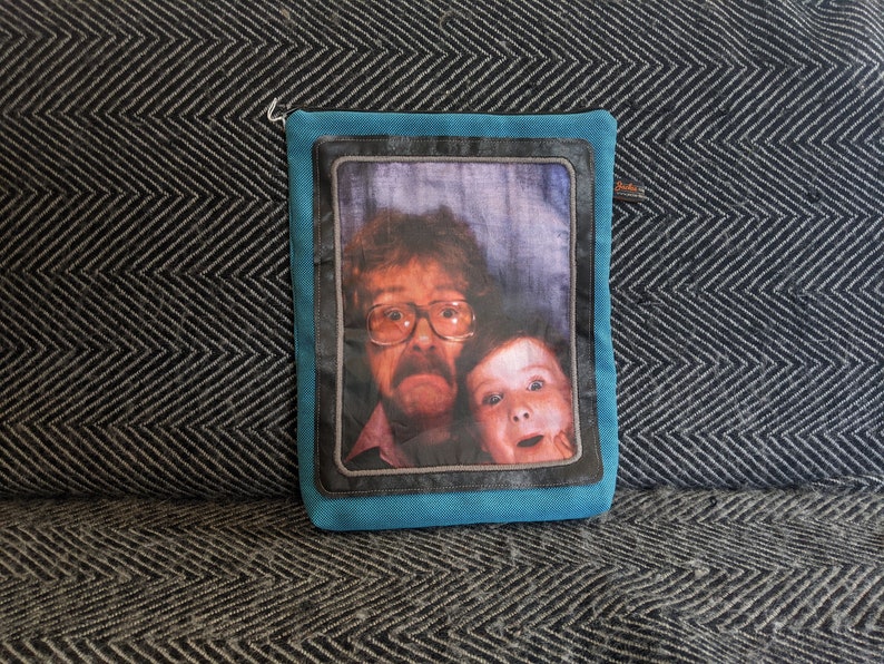 K&ouml;nnte beinhalten: Eine blaue Stofftasche mit einem schwarzen Rahmen und einem Foto von zwei Personen darin. Das Foto zeigt einen Mann mit Brille und Bart und ein kleines Kind mit einem &uuml;berraschten Ausdruck.
