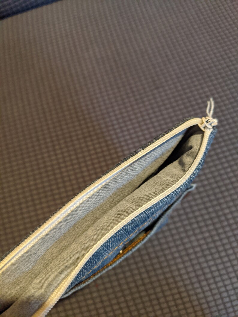 May include: A blue denim pencil case with a white zipper and a white lining. The case is open and the lining is visible.