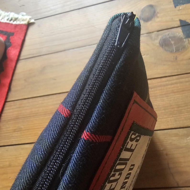 May include: Close-up of a zippered case with a plaid pattern in navy blue, red, and green. The case has a black zipper and a rectangular label with text. The case is likely for storing items.