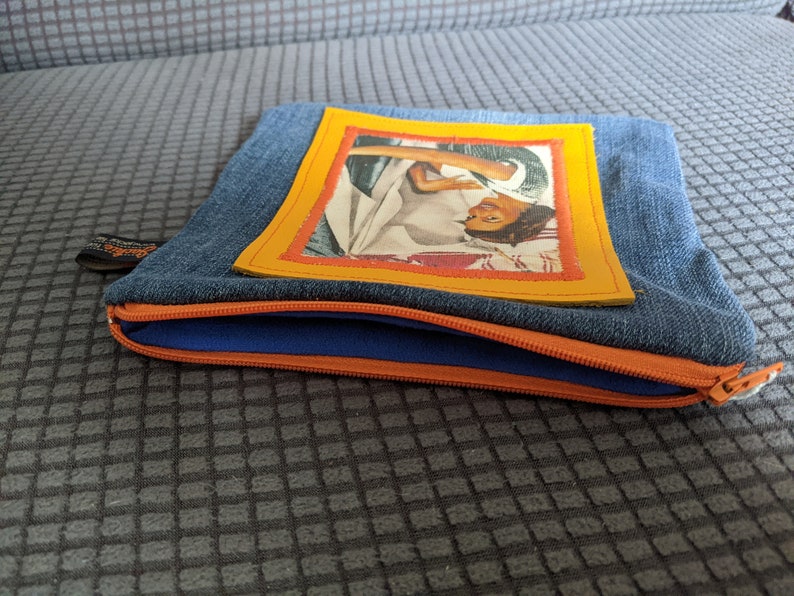 May include: A blue denim zippered pouch with a yellow frame around a vintage photo of a woman. The pouch has a blue zipper and an orange zipper pull.