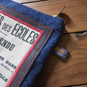 May include: Close-up of a denim pouch with a cream-colored patch featuring French text and a red stitched border. The pouch has a gold zipper and a small tag that reads "Jackie Fieur".