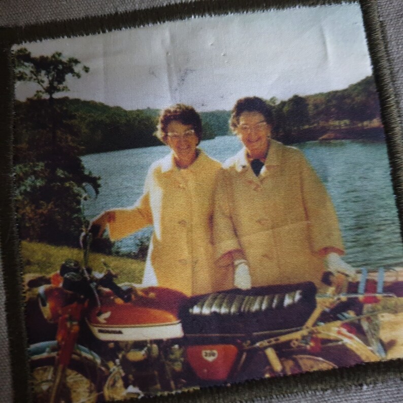 May include: Two women in yellow coats stand in front of a lake with a red and black motorcycle in the foreground.