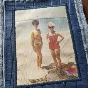 K&ouml;nnte beinhalten: Ein Vintage-Foto von zwei Frauen in Badeanz&uuml;gen, die an einem Strand stehen. Das Foto ist in einem blauen lederartigen Material gerahmt und auf einem grauen, strukturierten Stoff montiert.