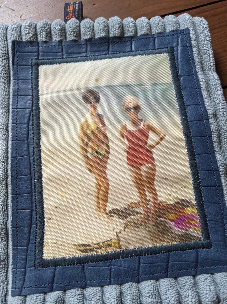 K&ouml;nnte beinhalten: Ein Vintage-Foto von zwei Frauen in Badeanz&uuml;gen, die an einem Strand stehen. Das Foto ist in einem blauen lederartigen Material gerahmt und auf einem grauen, strukturierten Stoff montiert.