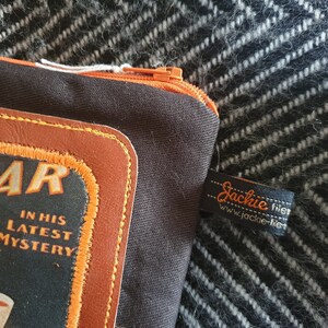 Pu&ograve; includere: Una pochette nera in tela con zip e una toppa in pelle marrone che dice "AR IN HIS LATEST MYSTERY". La pochette ha un'etichetta nera e arancione che dice "Jackie filer www.jackie-filer.com".