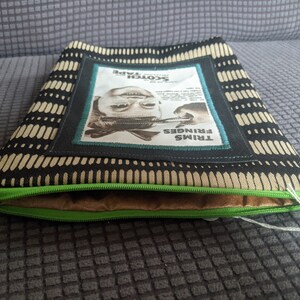 May include: A black and beige zippered pouch with a green zipper. The pouch has a rectangular patch with a black and white image of a person's face and the text "TRIMS FRINGES Scotch Tape".