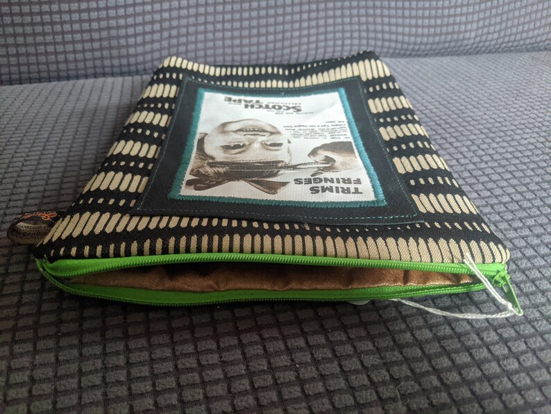May include: A black and beige zippered pouch with a green zipper. The pouch has a rectangular patch with a black and white image of a person's face and the text "TRIMS FRINGES Scotch Tape".