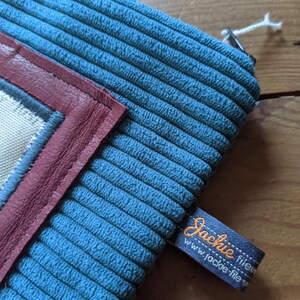 Peut inclure: Une pochette zipp&eacute;e en velours c&ocirc;tel&eacute; bleu avec un rabat en cuir bordeaux et un insert en tissu blanc. La pochette porte une &eacute;tiquette noire avec le texte "Jackie fileu" et "www.jackie-fileu.com" en orange.