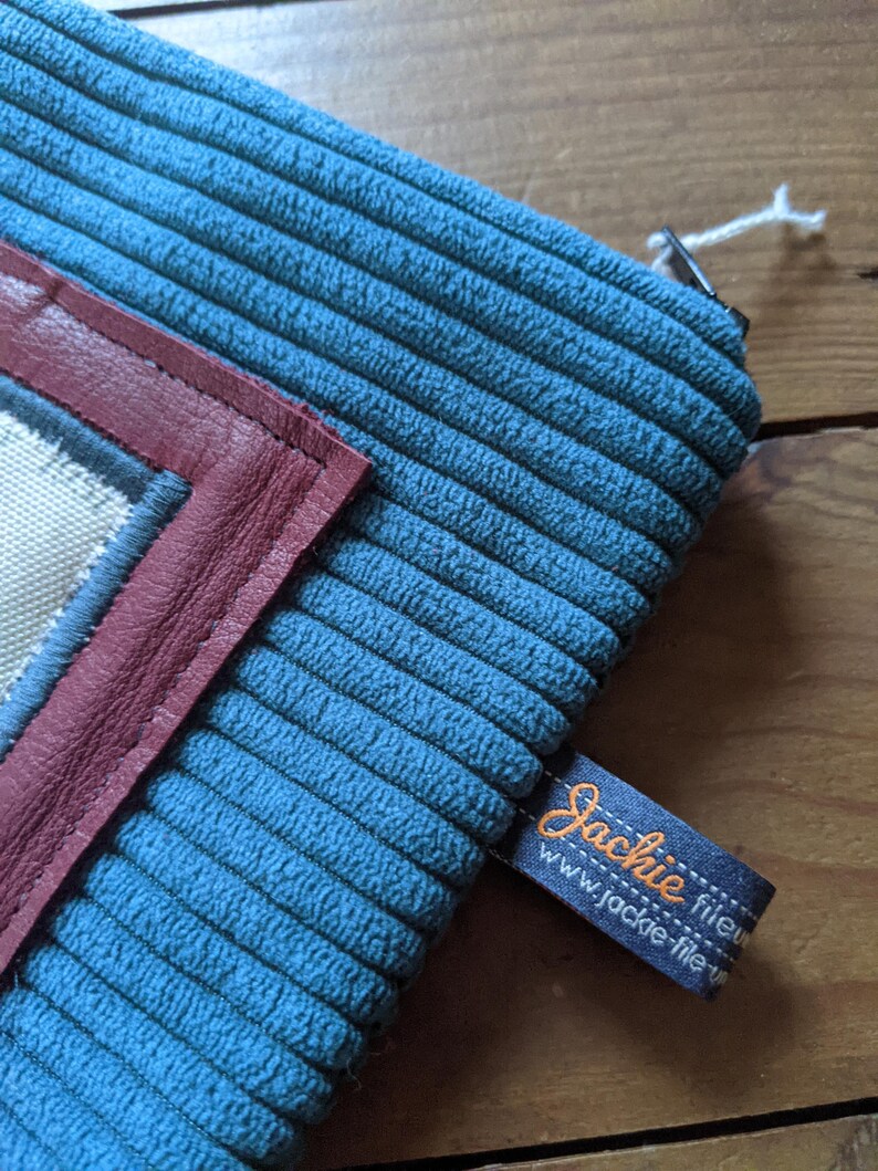 Peut inclure: Une pochette zipp&eacute;e en velours c&ocirc;tel&eacute; bleu avec un rabat en cuir bordeaux et un insert en tissu blanc. La pochette porte une &eacute;tiquette noire avec le texte "Jackie fileu" et "www.jackie-fileu.com" en orange.